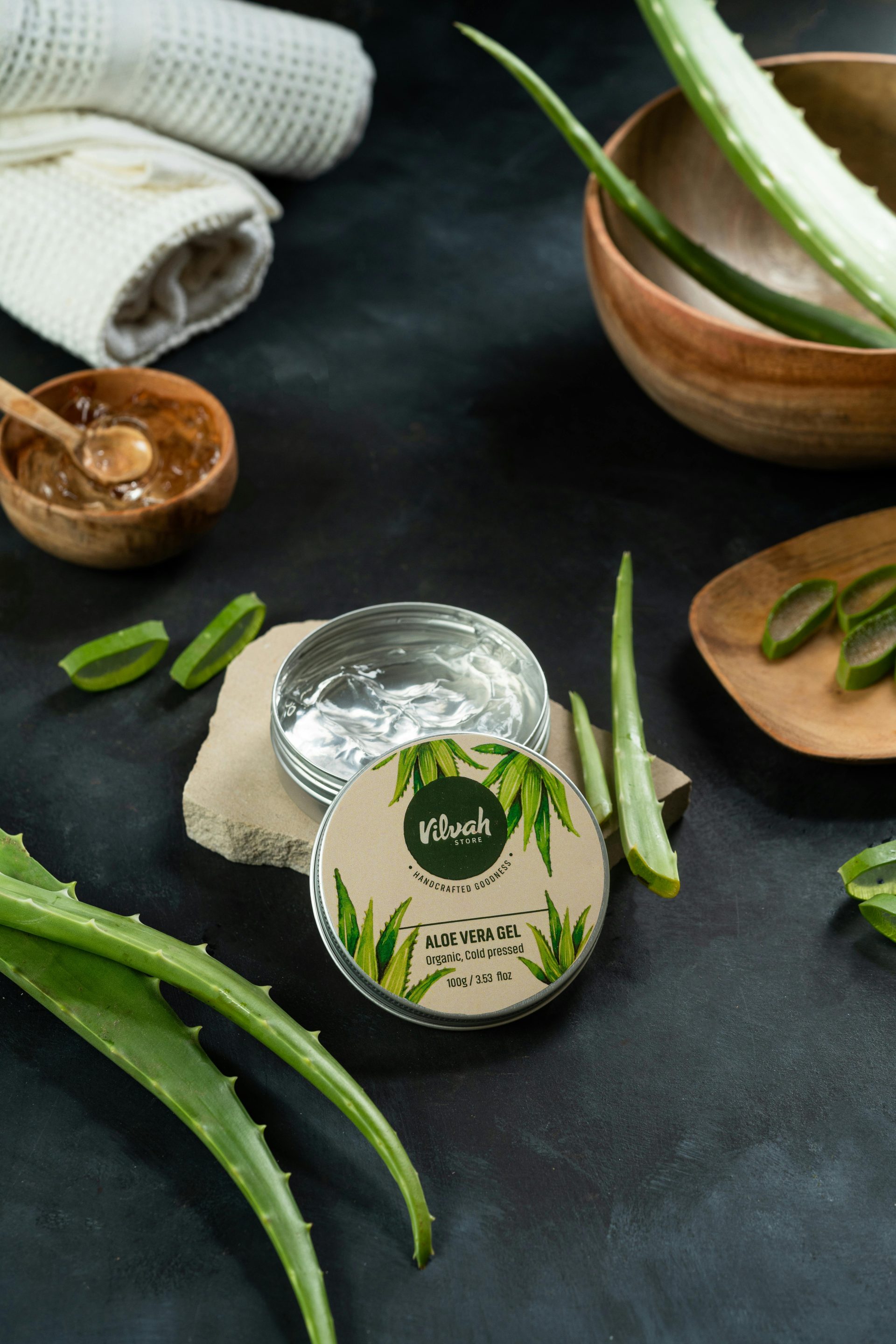 Aloe vera gel jar surrounded by fresh aloe leaves and spa items on a dark surface.
